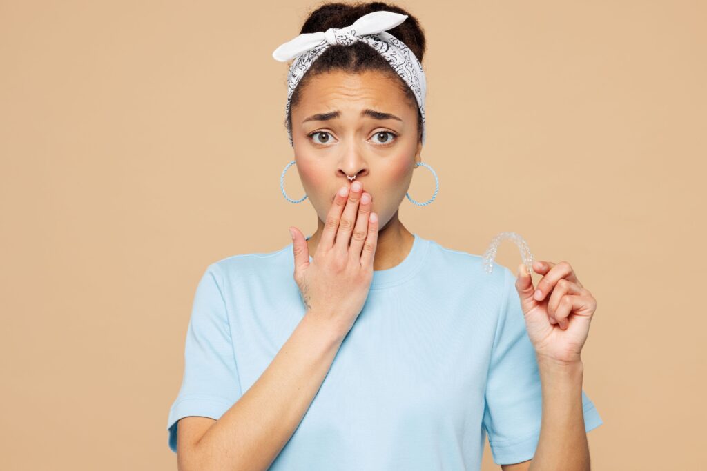 Woman in blue t-shirt holding Invisalign and covering mouth in "oh-no!" expression